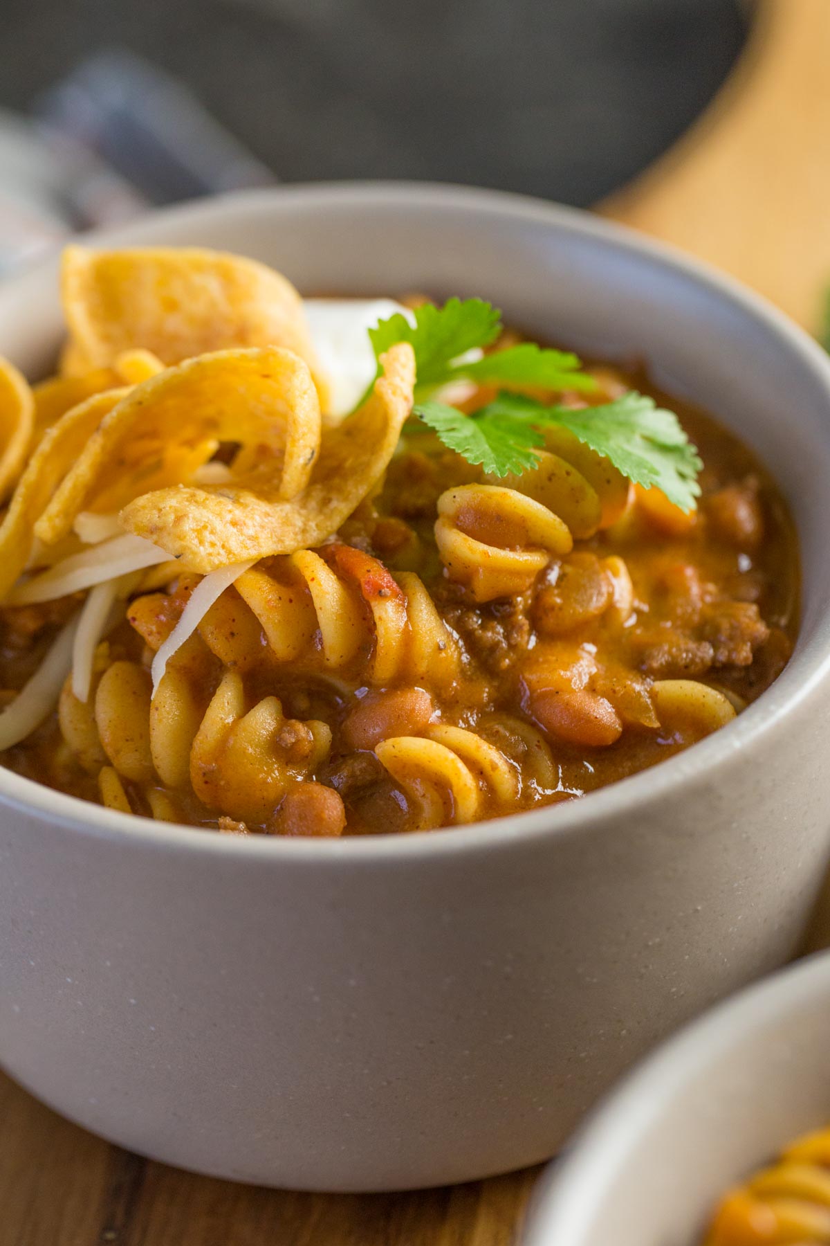 A close up shot of a grey bowl filled with Beef Queso Chili with pasta, Fritos, source cream, and cilantro. 