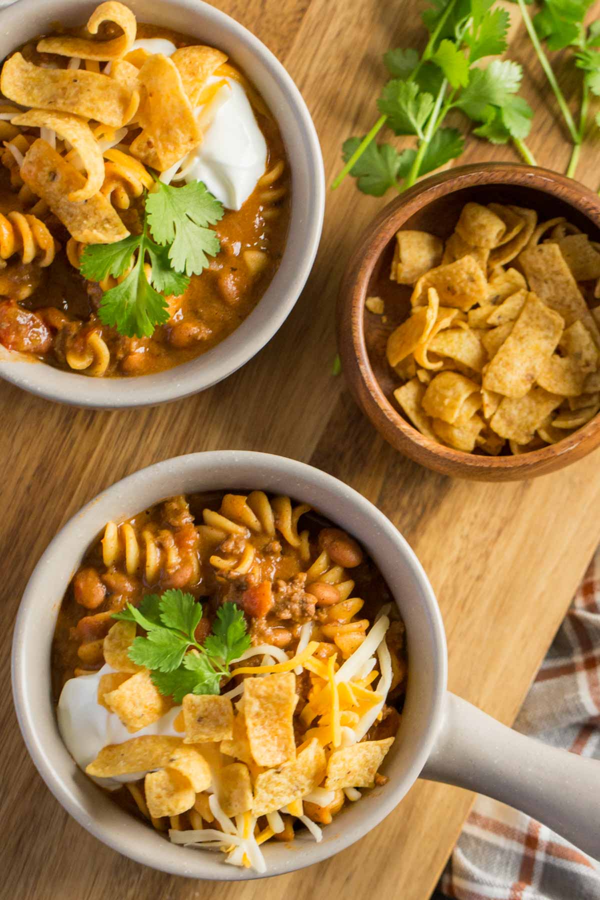 A top down view of two bowls Beef Queso Chili topped with sour cream, Fritos and Cilantro on a wooden board. 