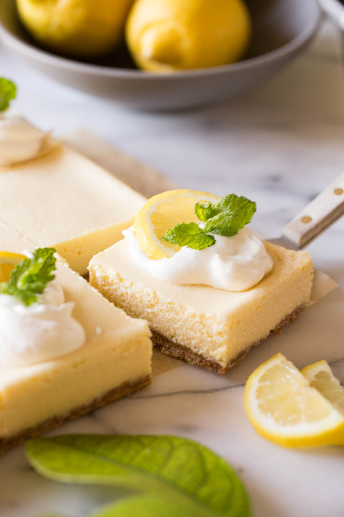 Close up view of Best Creamy Lemon Bar being lifted out of the pan with fresh lemons in the background.