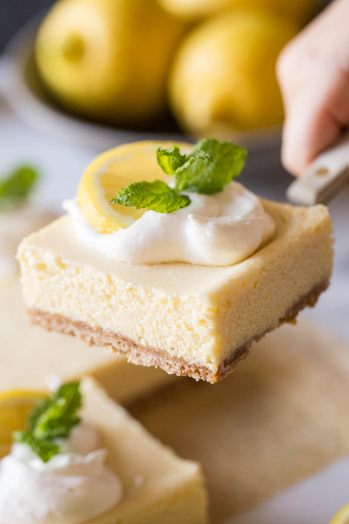 Close up view of Best Creamy Lemon Bar being lifted out of the pan with fresh lemons in the background.