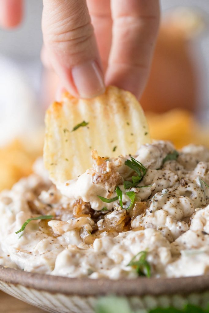 Chip dipping into Bowl of Roasted Garlic and Caramelized Onion Dip on a platter with chips. 