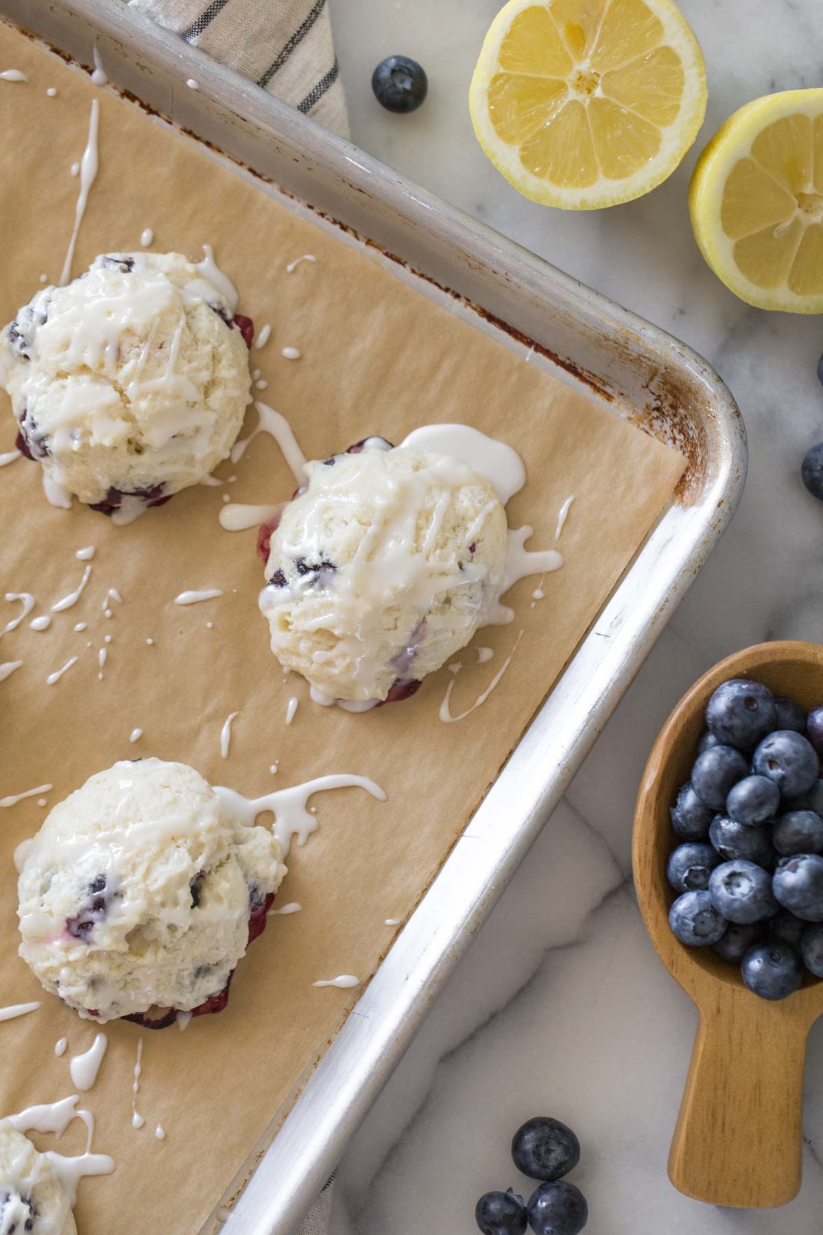 Glazed Lemon Blueberry Muffin Tops on a baking sheet with a grey background with fresh lemons and blueberries. 