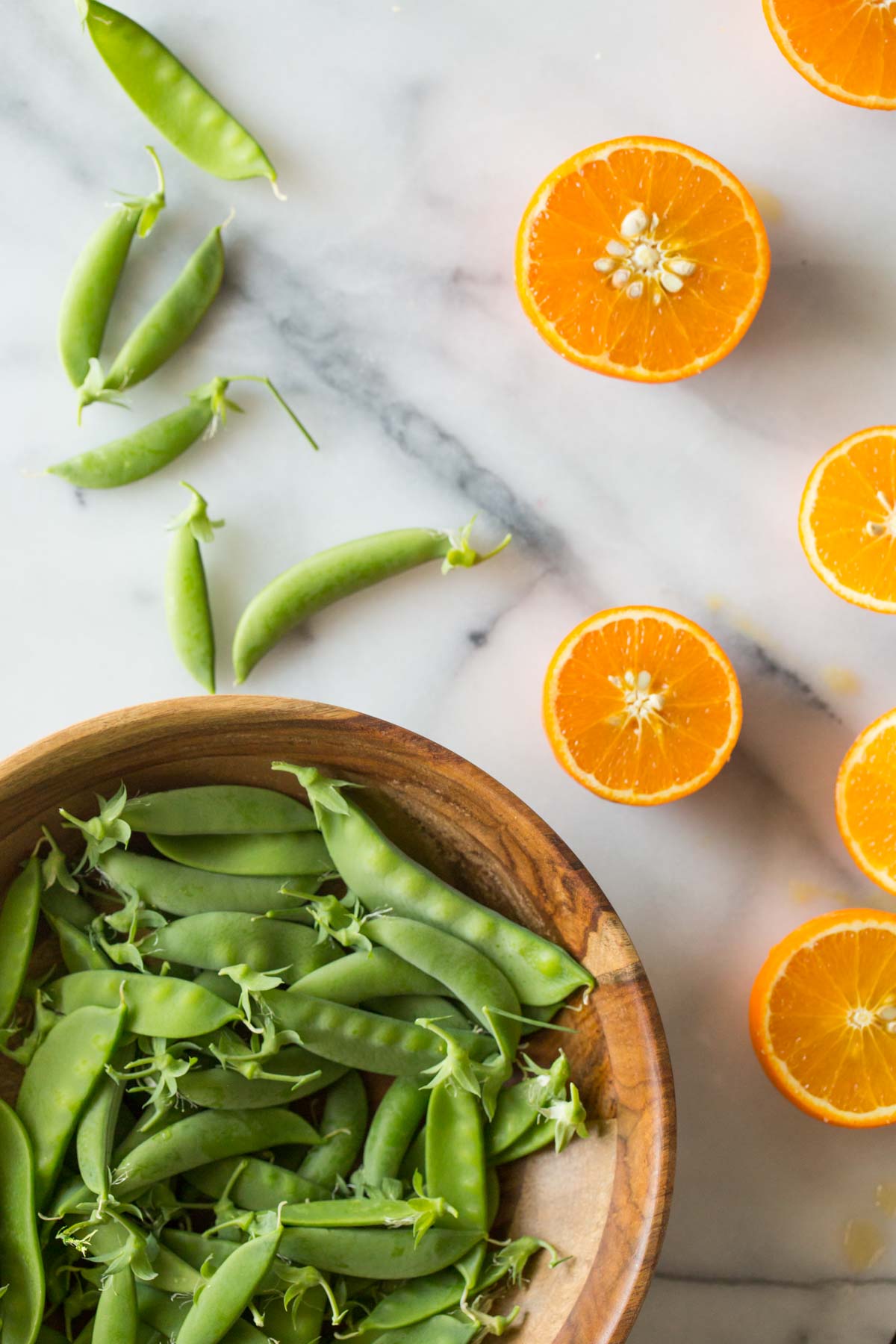 A flat lay overhead shot of sliced oranges and sugar snap peas on a marble background. 