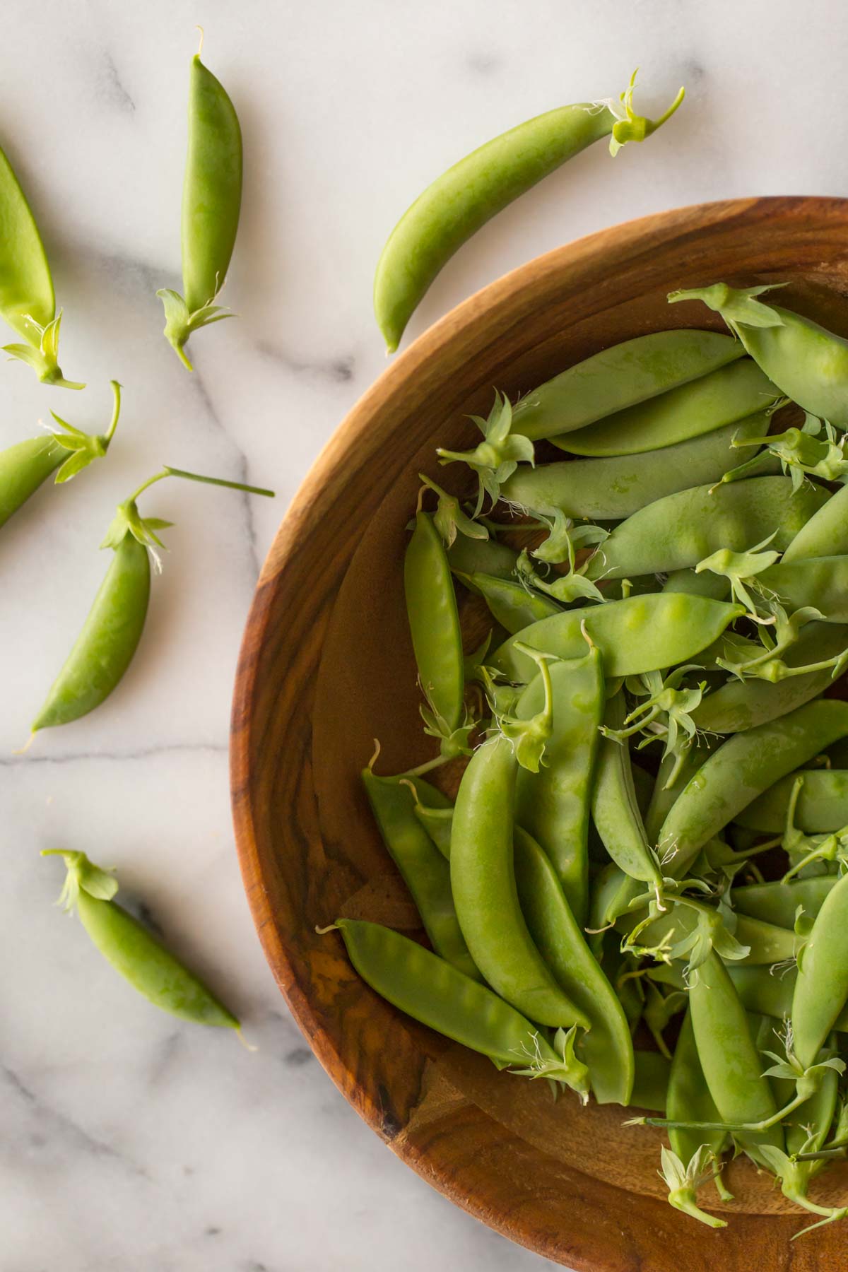 Overhead shot of a wooden bowl full of sugar snap peas on a white marble background. 
