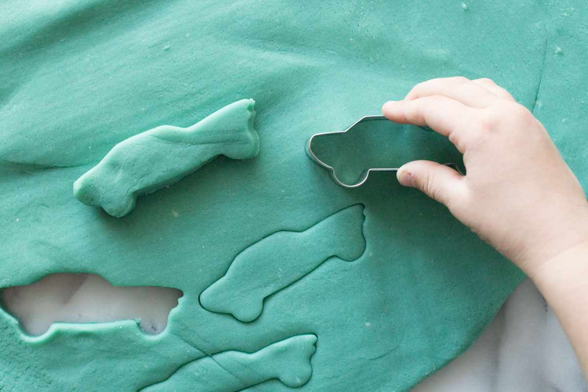 Overhead view of blue playdough with little hands using a car shaped cutter. 