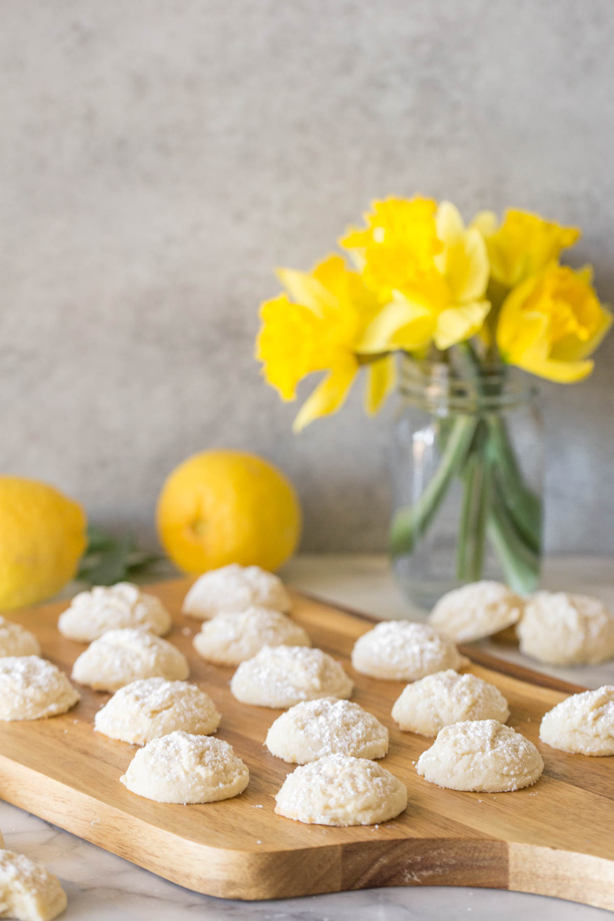 Lemon Cookies on a wooden cutting board with yellow daffodils in the background. 