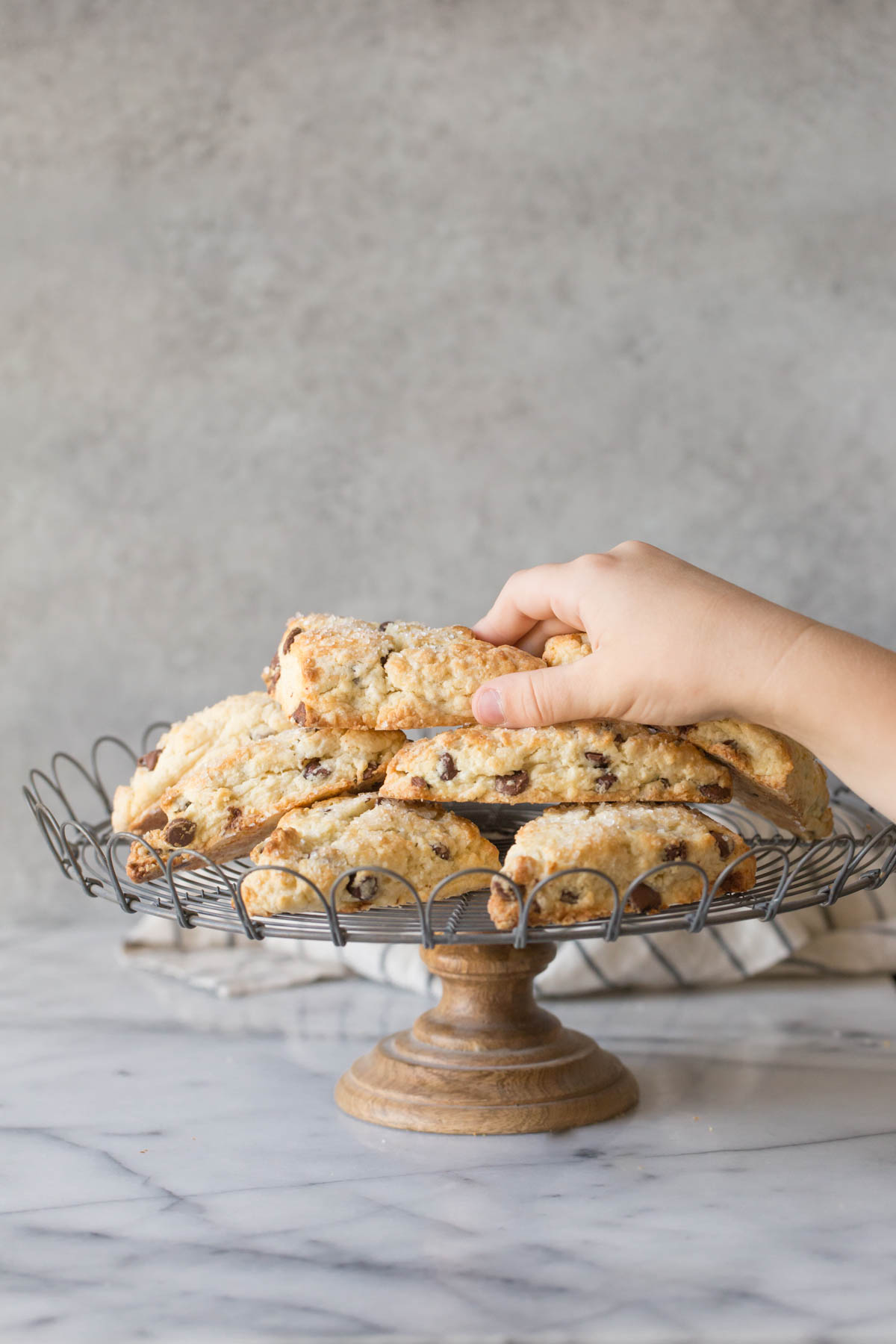 A stack of Chocolate Chip Scones on a cake stand with a hand reaching for the scone on top.  