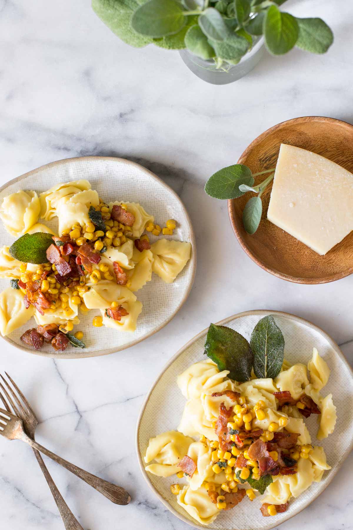 Overhead view of two plates of Creamy Tortellini With Bacon and Corn and a wooden plate with a block of Parmesan cheese, on a marble background.  