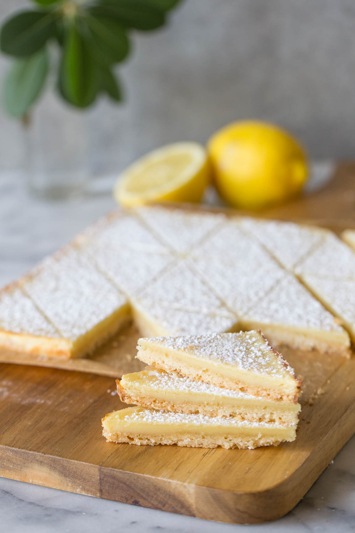 Three Swedish Lemon Bars stacked on a wooden cutting board, with more bars and lemons in the background.  
