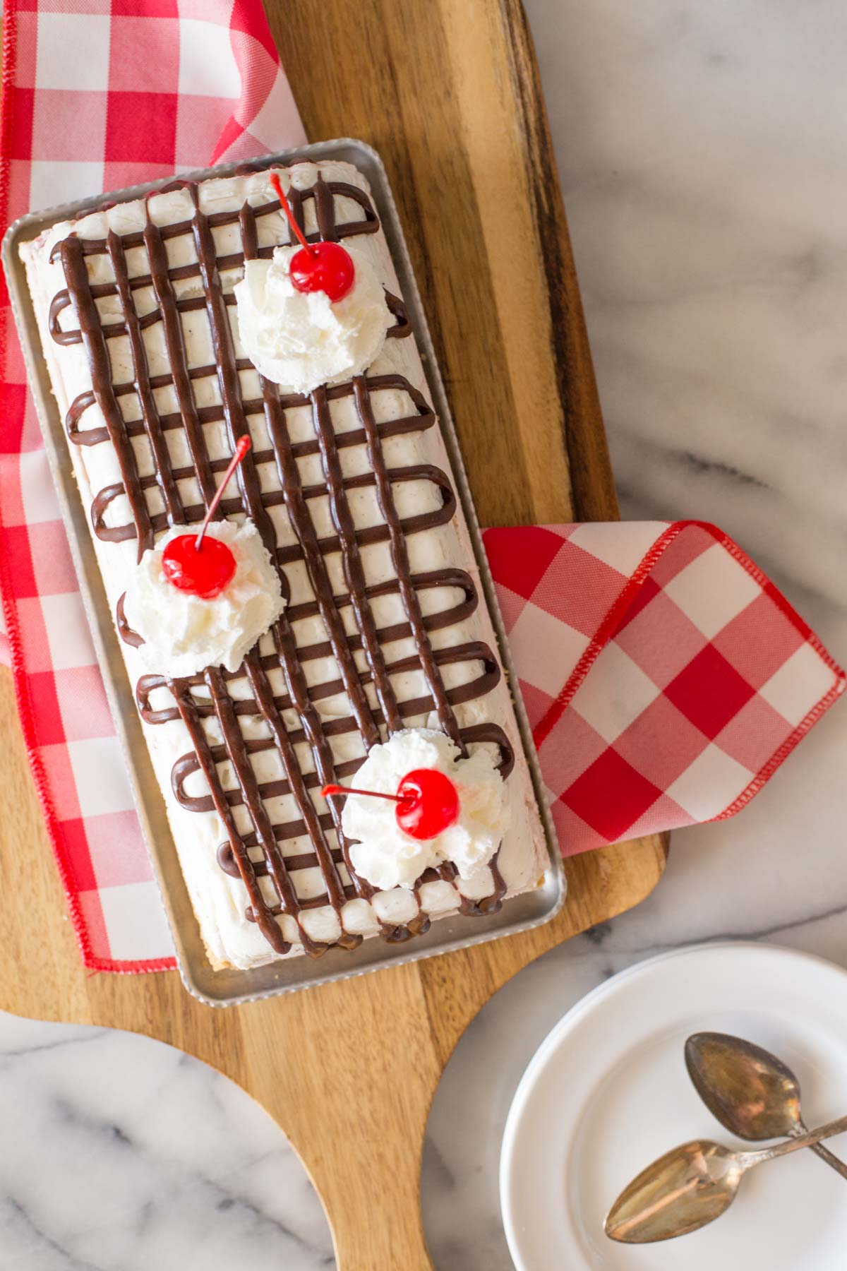 Overhead shot of Banana Split Ice Cream Cake on a serving tray sitting on a red and white checkered cloth on a wooden cutting board.  
