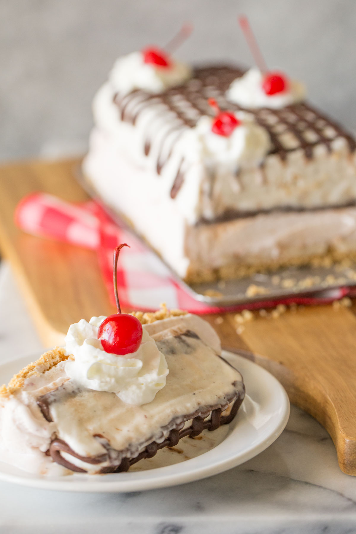 A slice of Banana Split Ice Cream Cake on a plate topped with whipped cream and a cherry, with the rest of the Banana Split Ice Cream Cake in the background. 