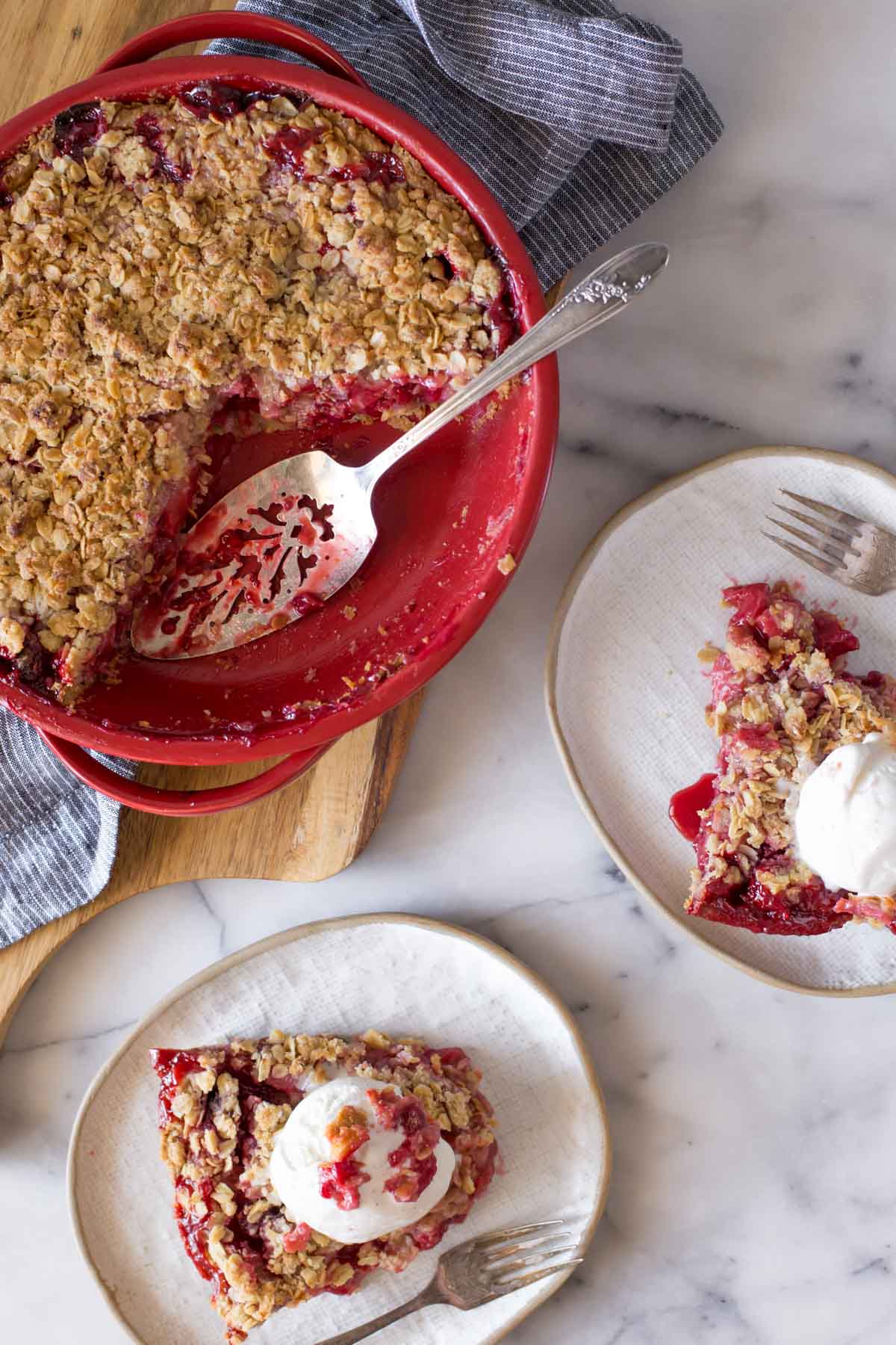 Overhead shot of Berry Crumble Pie in a pie dish with a serving utensil, sitting next to two plates with slices of Berry Crumble Pie each topped with a scoop of vanilla ice cream.  