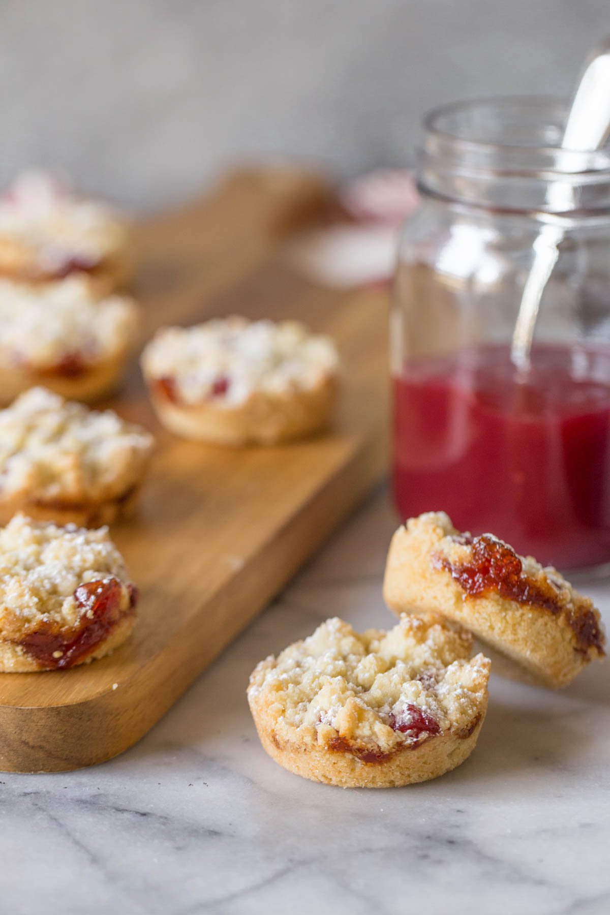 Two Costco Raspberry Crumble Cookies with a jar of raspberry preserves in the background, along with more cookies on a wooden cutting board. 