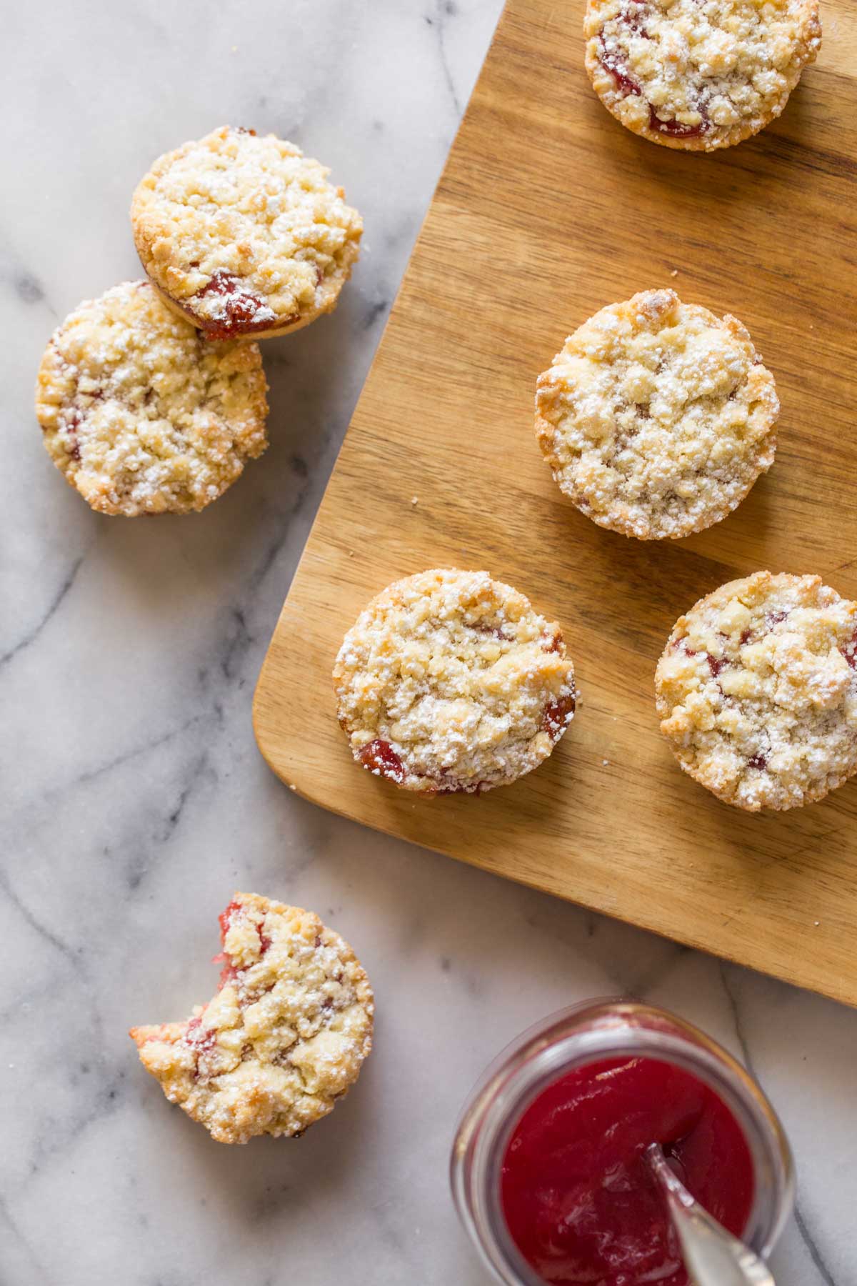 Overhead shot of Costco Raspberry Crumble Cookies on a wooden cutting board, with a few cookies next to the cutting board along with a jar of raspberry preserves.  