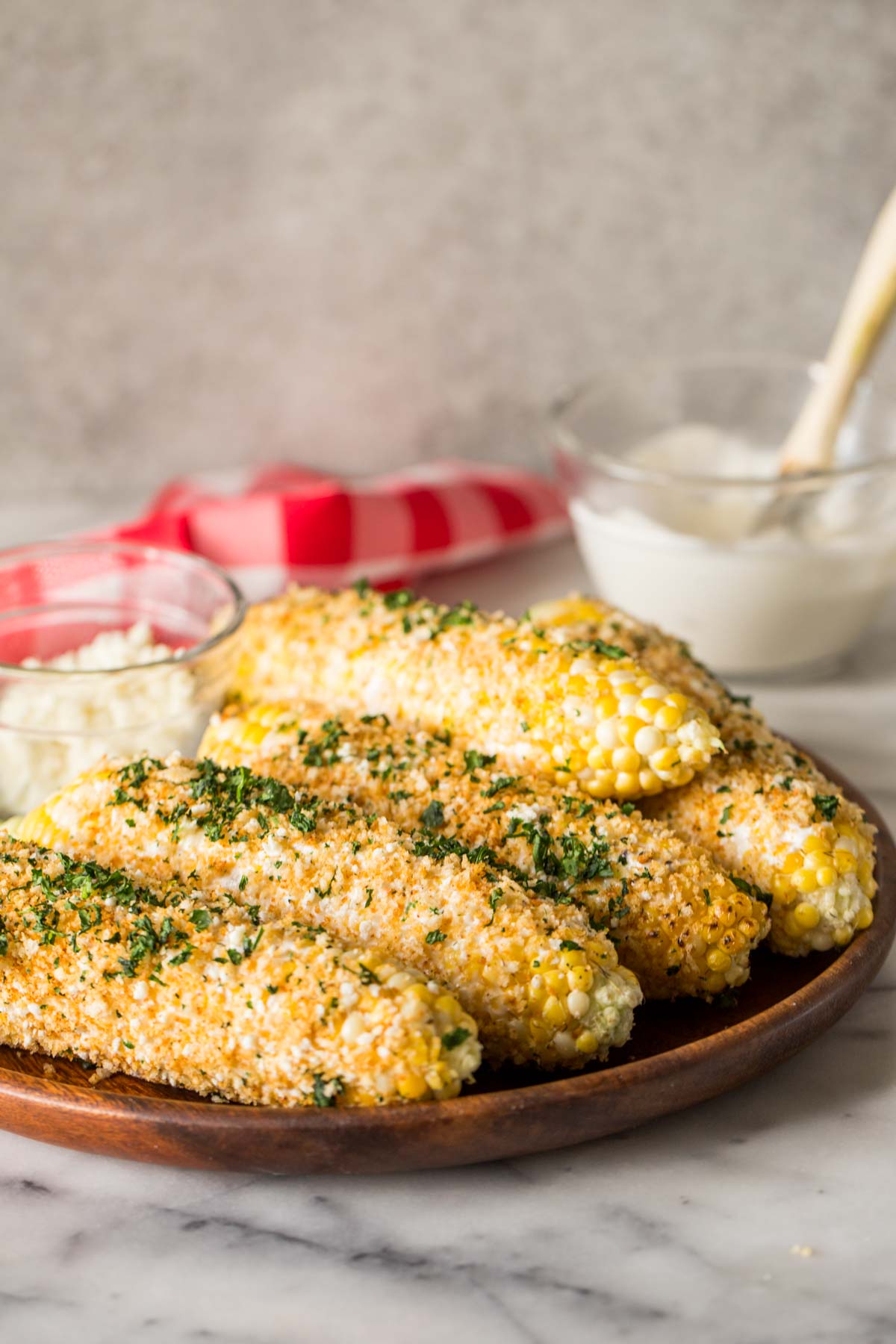 Mexican Street Corn With Panko and a small glass dish of cotija cheese on a wood platter, with a glass bowl of the mayonnaise mixture in the background. 