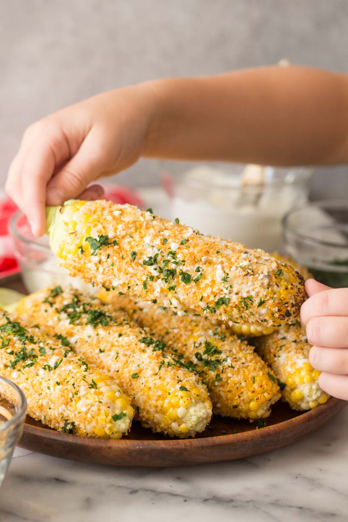 Two hands holding an ear of Mexican Street Corn With Panko, with more ears of corn on a wood platter in the background. 