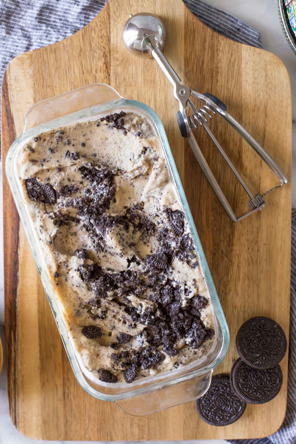 Overhead shot of Peanut Butter Oreo Ice Cream in a glass dish, sitting on a wooden cutting board with an ice cream scoop and Oreo cookies. 