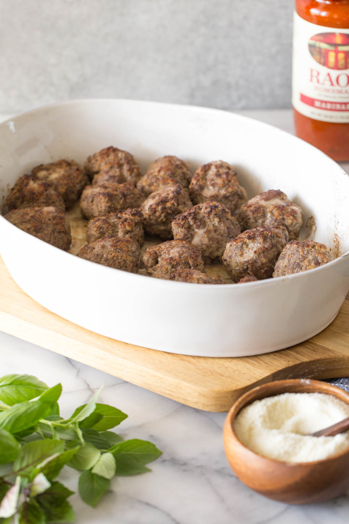 Easy Broiled Italian Meatballs in a baking dish on a wooden cutting board, after being broiled but before the sauce is added, with a jar of pasta sauce and a small wood bowl of Parmesan cheese both next to the cutting board. 