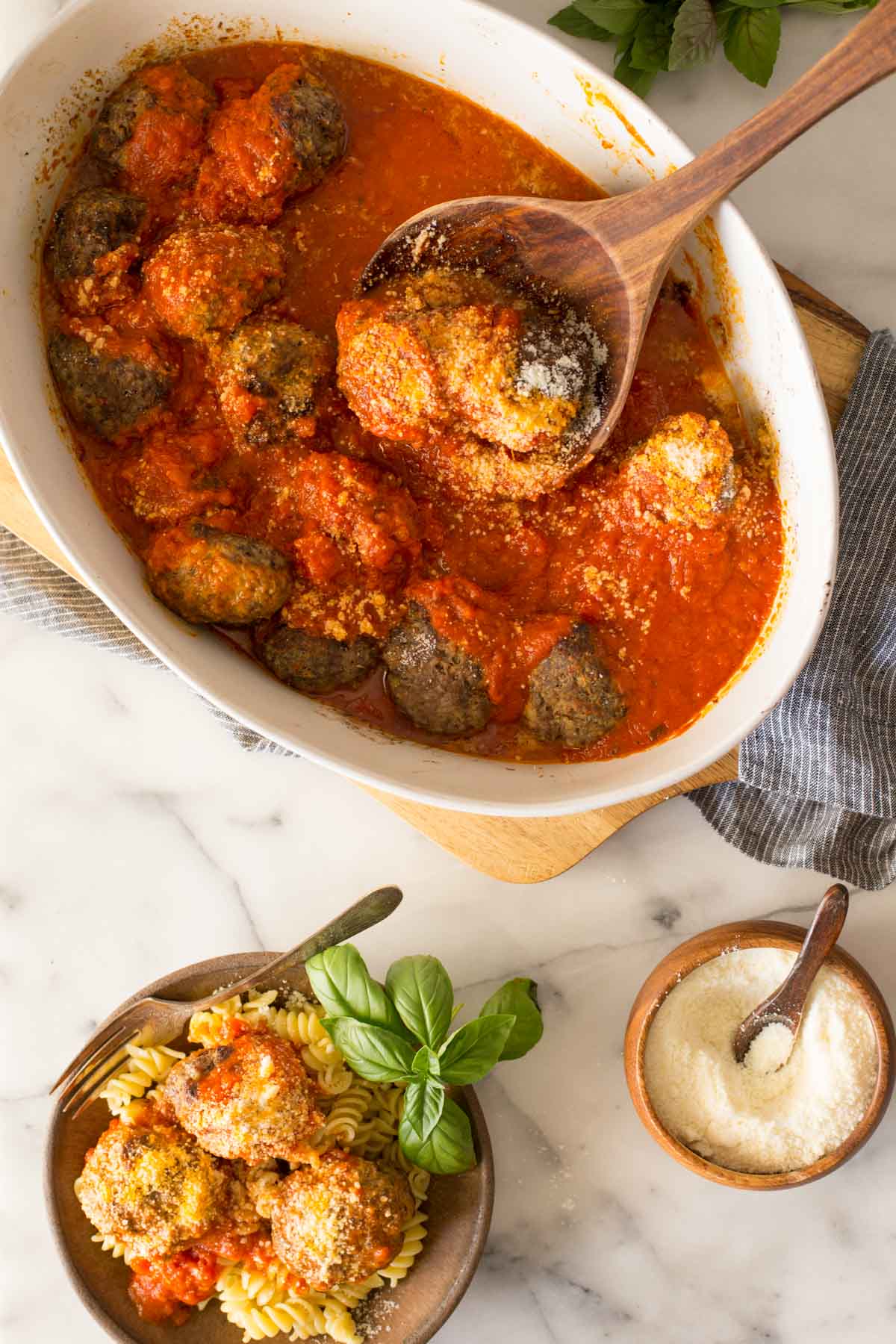 Overhead shot of Easy Broiled Italian Meatballs in a baking dish with a wood serving spoon in the meatballs, and a bowl of meatballs served over rotini pasta and a small wood bowl of Parmesan cheese both next to the cutting board.  