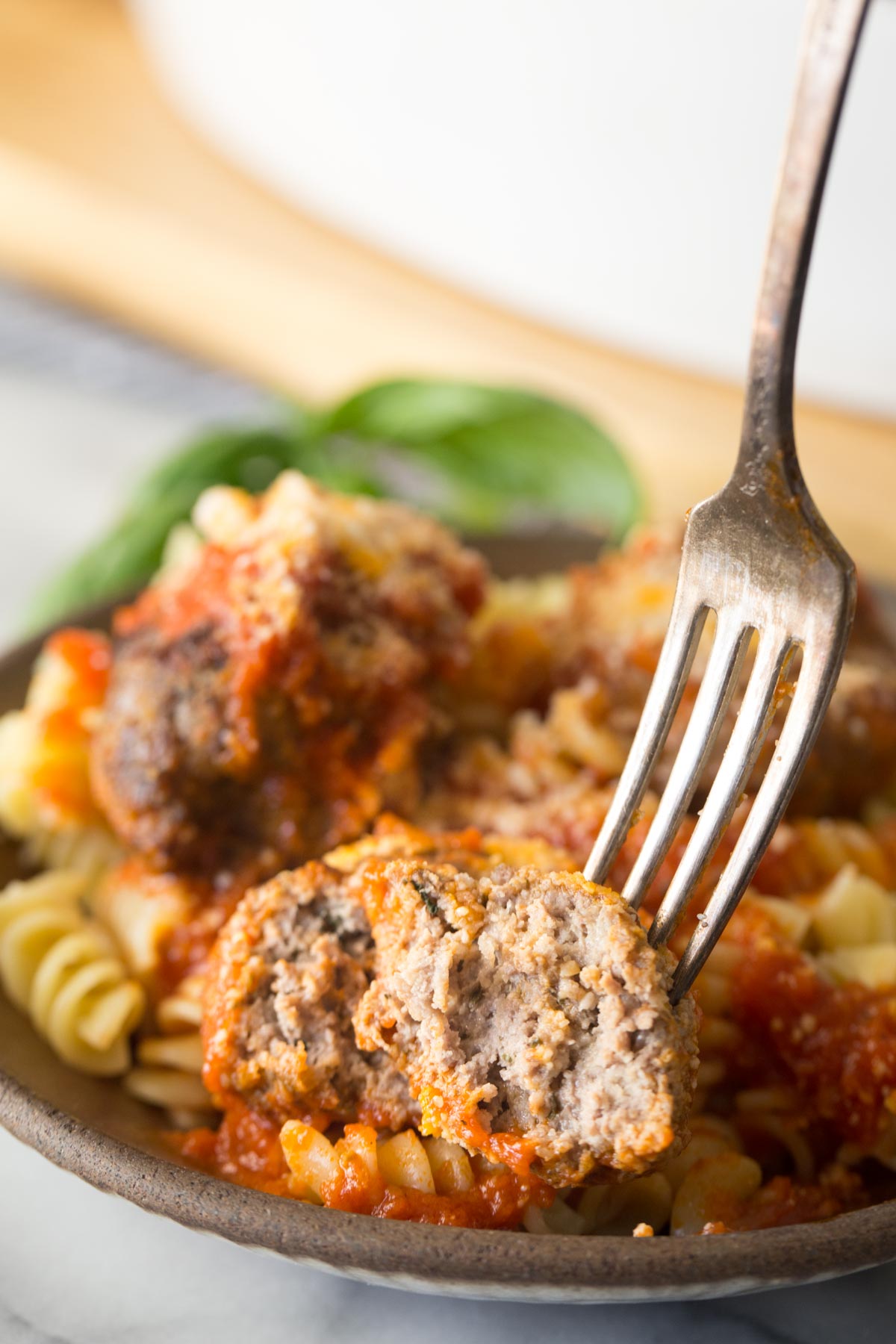 Close up shot of a fork in half of an Easy Broiled Italian Meatball, showing the inside texture, with a bowl of meatballs served over rotini pasta in the background. 