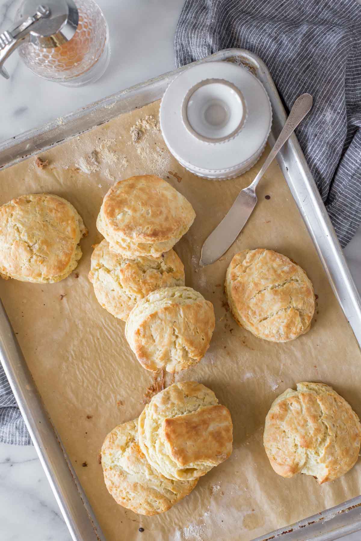 Overhead shot of Easy Homemade Biscuits, along with a butter dish and butter knife on a parchment lined baking sheet, sitting on a blue cloth next to a glass honey dispenser with a marble background. 