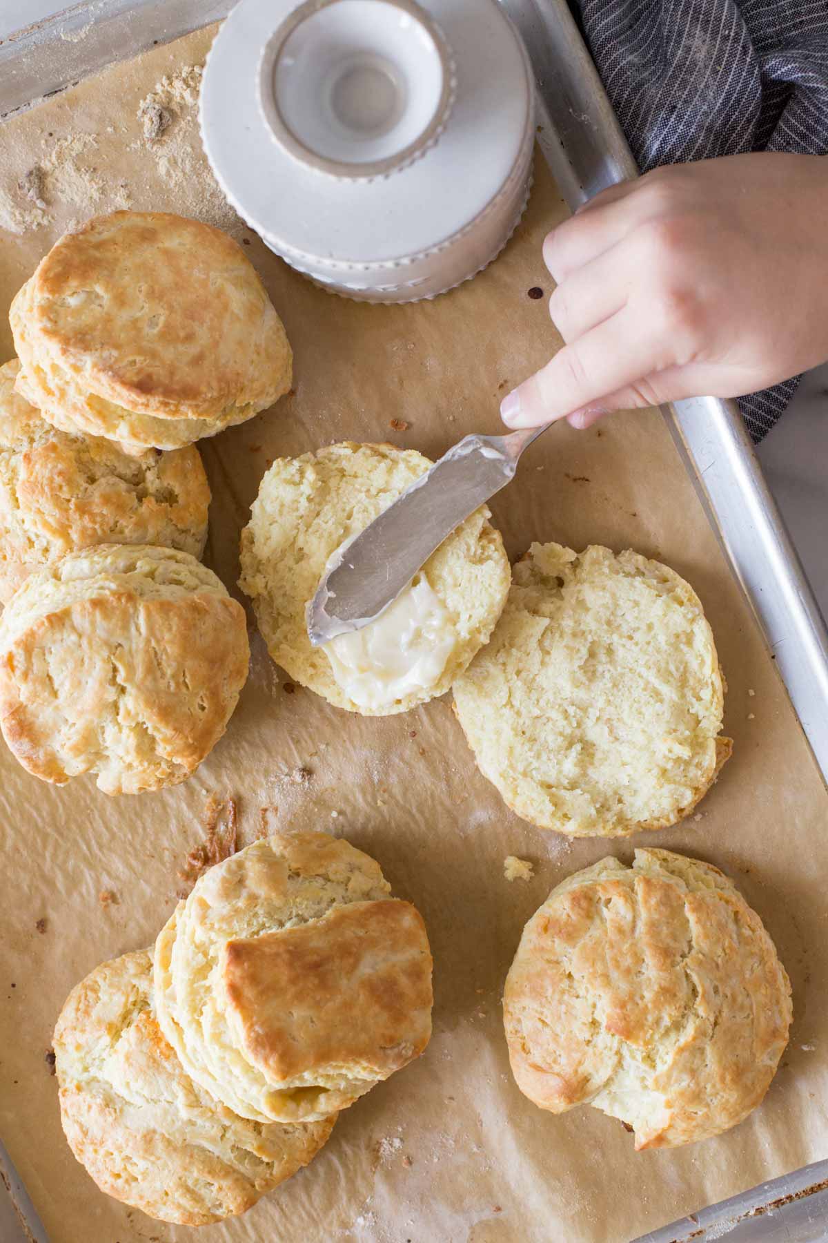 Overhead shot of Easy Homemade Biscuits and a butter dish on a parchment lined baking sheet, with a hand spreading butter on an open biscuit.  