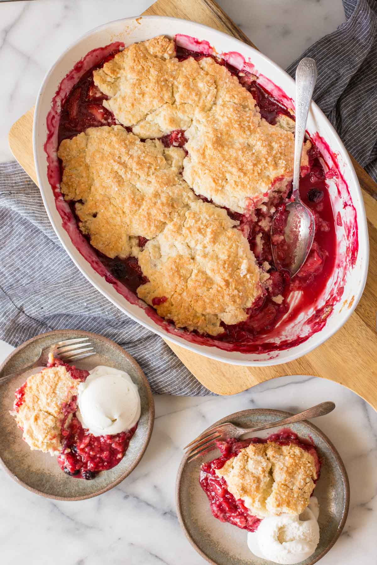 Overhead shot of Triple Berry Cobbler in a baking dish, with two plates of Triple Berry Cobbler each with a scoop of vanilla ice cream next to the baking dish.  
