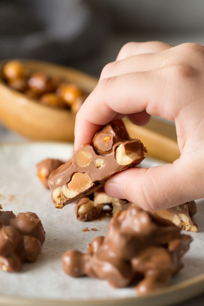 Close up shot of a hand holding a Chocolate Peanut Cluster that has been cut in half to show the inside of the cluster.  