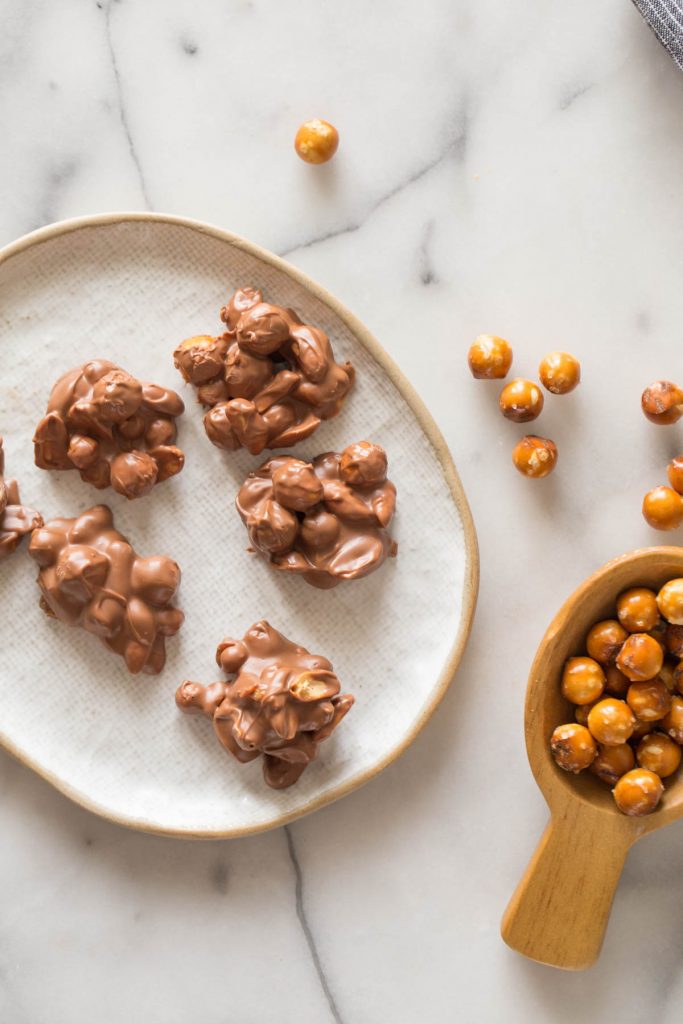 Overhead shot of Chocolate Peanut Clusters on a plate, with a wooden spoon full of pretzel rounds next to the plate. 