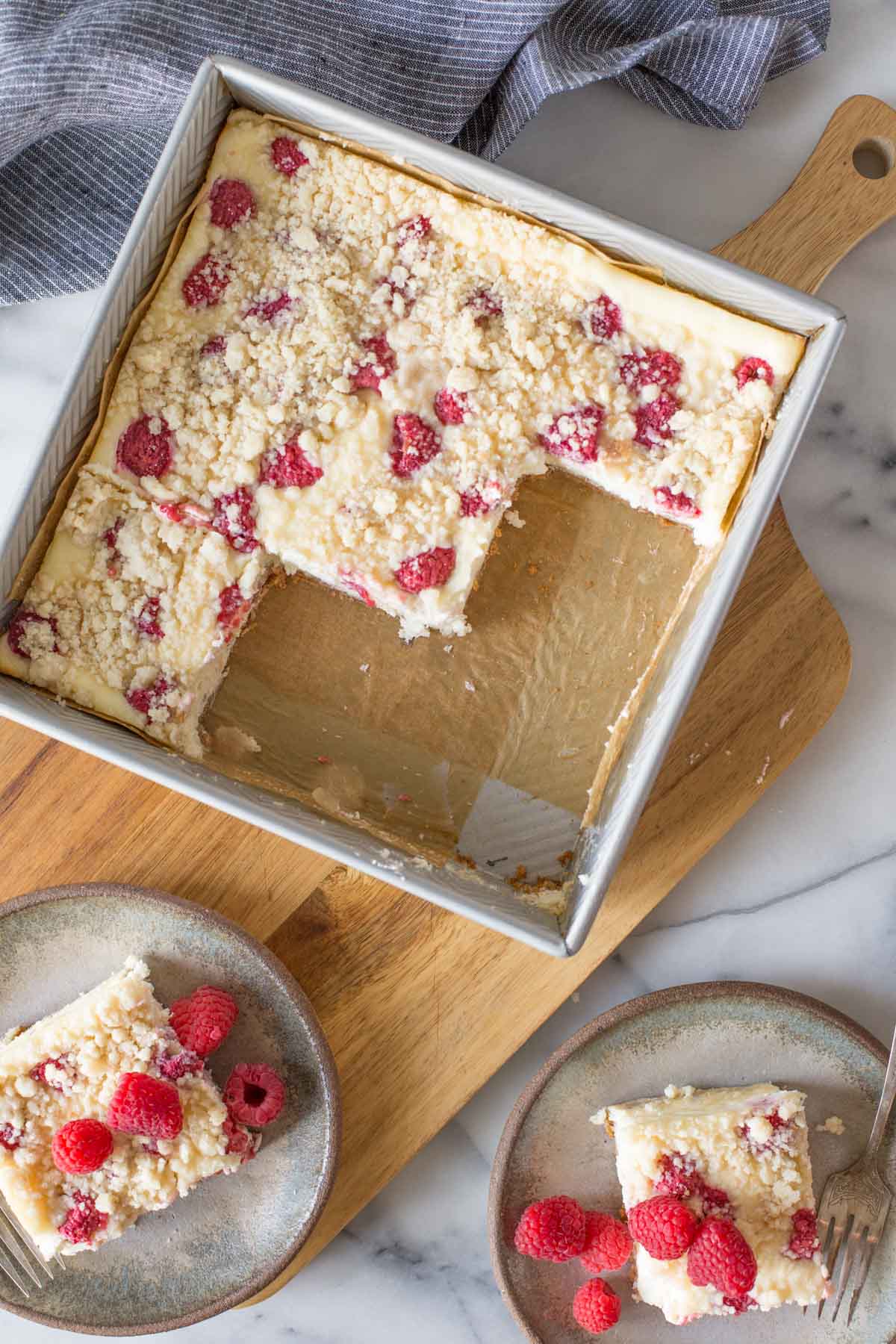 Overhead shot of a baking pan of Cheesecake Bars With Berry Streusel on a wooden cutting board, with two pieces served on plates with fresh raspberries next to it.  