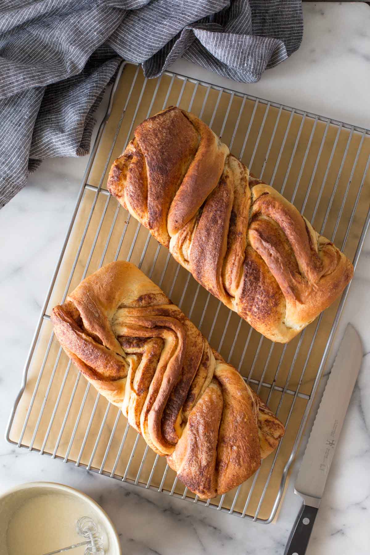 Overhead shot of two loaves of Cinnamon Twist Bread on a cooling rack with parchment paper under the rack, on a marble background.  