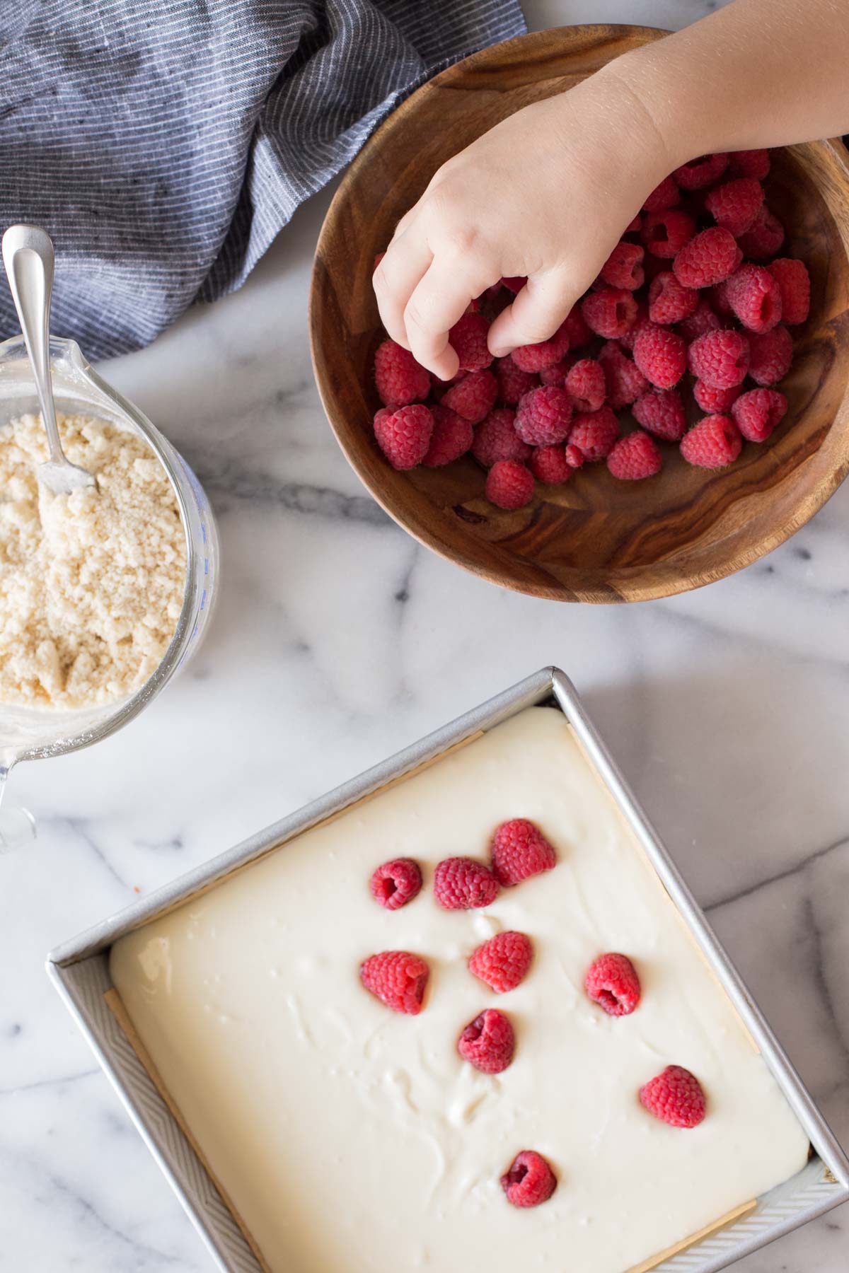 Overhead shot of a baking pan with the cheesecake being topped with fresh raspberries from a wooden bowl, and a glass measuring cup with the streusel topping next to it for the Cheesecake Bars With Berry Streusel.
