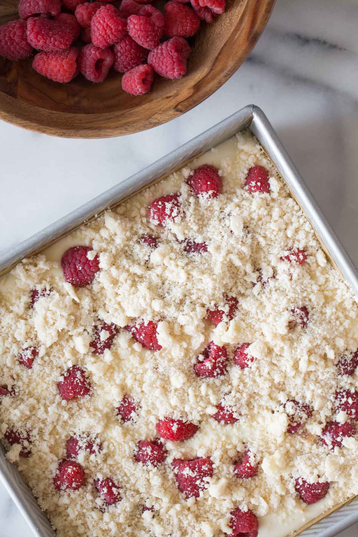 Overhead shot of Cheesecake Bars With Berry Streusel in a baking pan, next to a wooden bowl of fresh raspberries. 