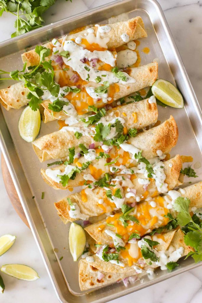Overhead shot of BBQ Chicken Taquitos on a baking sheet, garnished with lime wedges and fresh cilantro. 