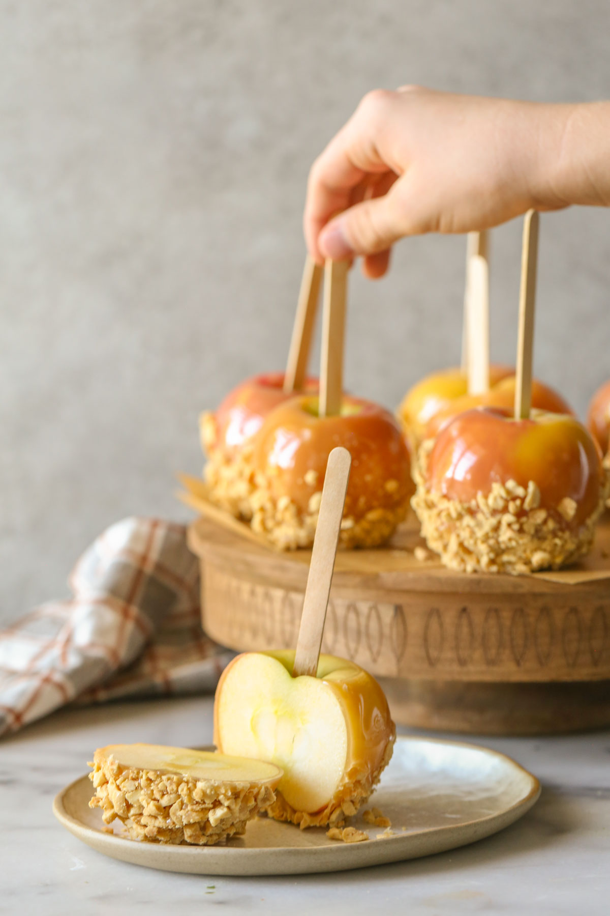 A Homemade Caramel Apple on a plate that has been cut in half, with more Homemade Caramel Apples on a wood stand in the background.  