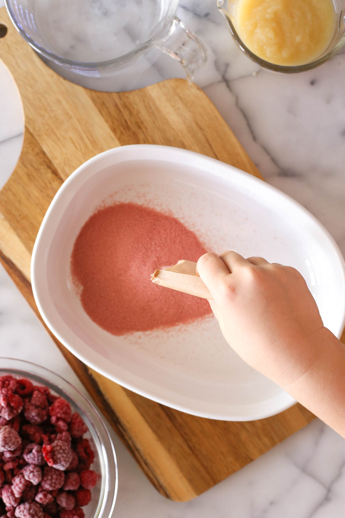Overhead shot of a package of raspberry gelatin being poured into a baking dish sitting on a wood cutting board, with a measuring cup of applesauce, a measuring cup of water and a bowl of frozen raspberries next to the cutting board.  