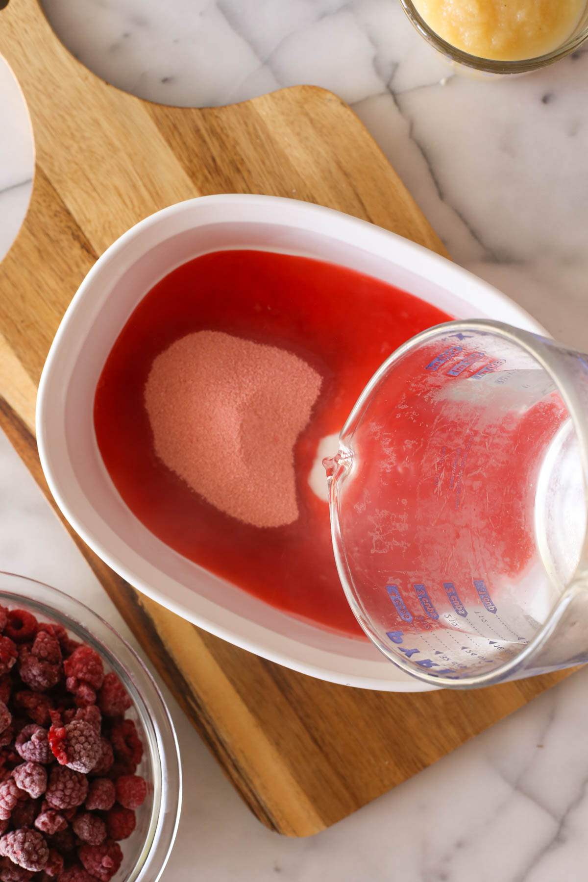 Overhead shot of a glass measuring cup of boiling water being poured in the baking dish with the raspberry gelatin, with a bowl of frozen raspberries next to it.  