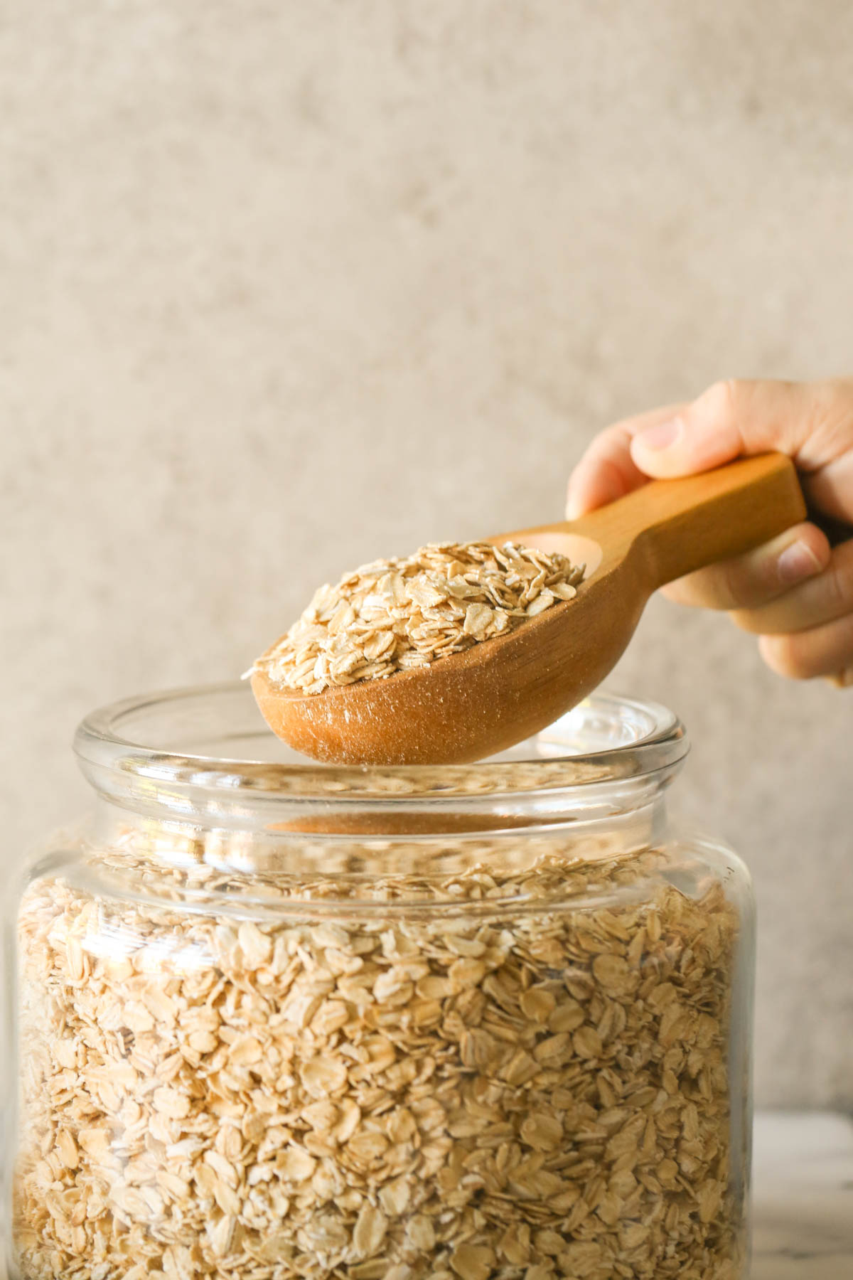 A large wooden scoop full of oats being held above a large glass jar of old fashioned oats.  