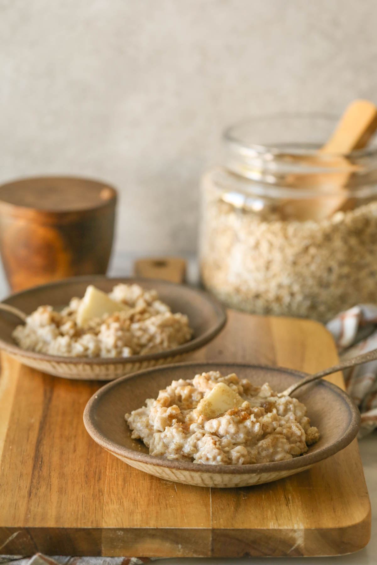 Two bowls of oatmeal topped with butter and brown sugar on a wood cutting board, with a wood salt container and a glass jar of oats in the background. 