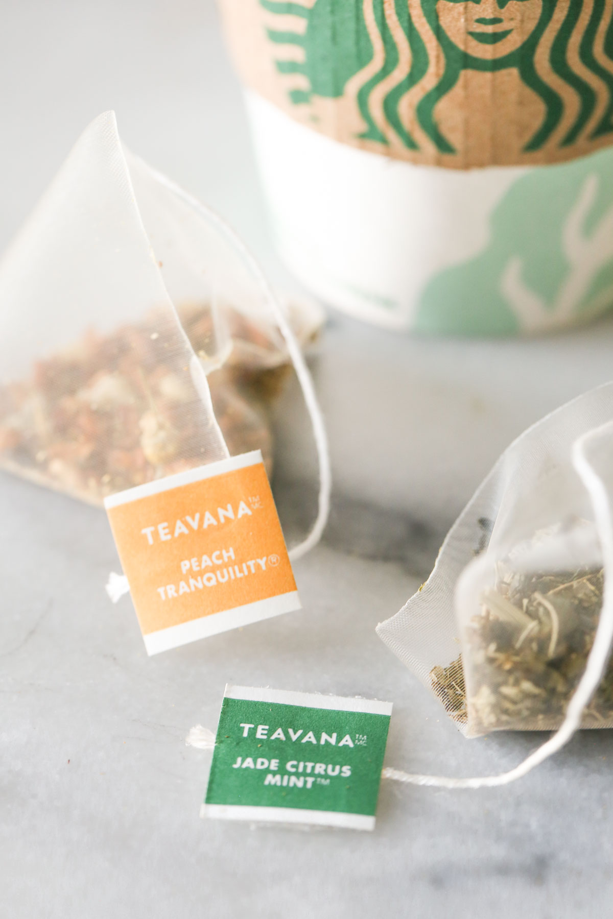 Close up shot of a Teavana Peach Tranquility tea sachet next to a Teavana Jade Citrus Mint tea sachet, with a Starbucks cup in the background. 