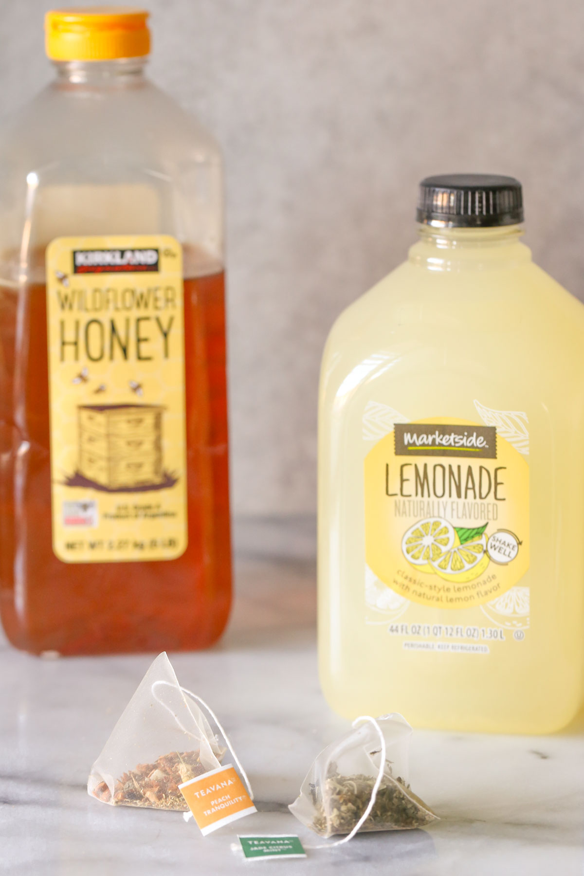 A Teavana Peach Tranquility tea sachet next to a Teavana Jade Citrus Mint tea sachet, with a jug of lemonade and a container of honey in the background, all ingredients for the Starbucks Medicine Ball Tea.