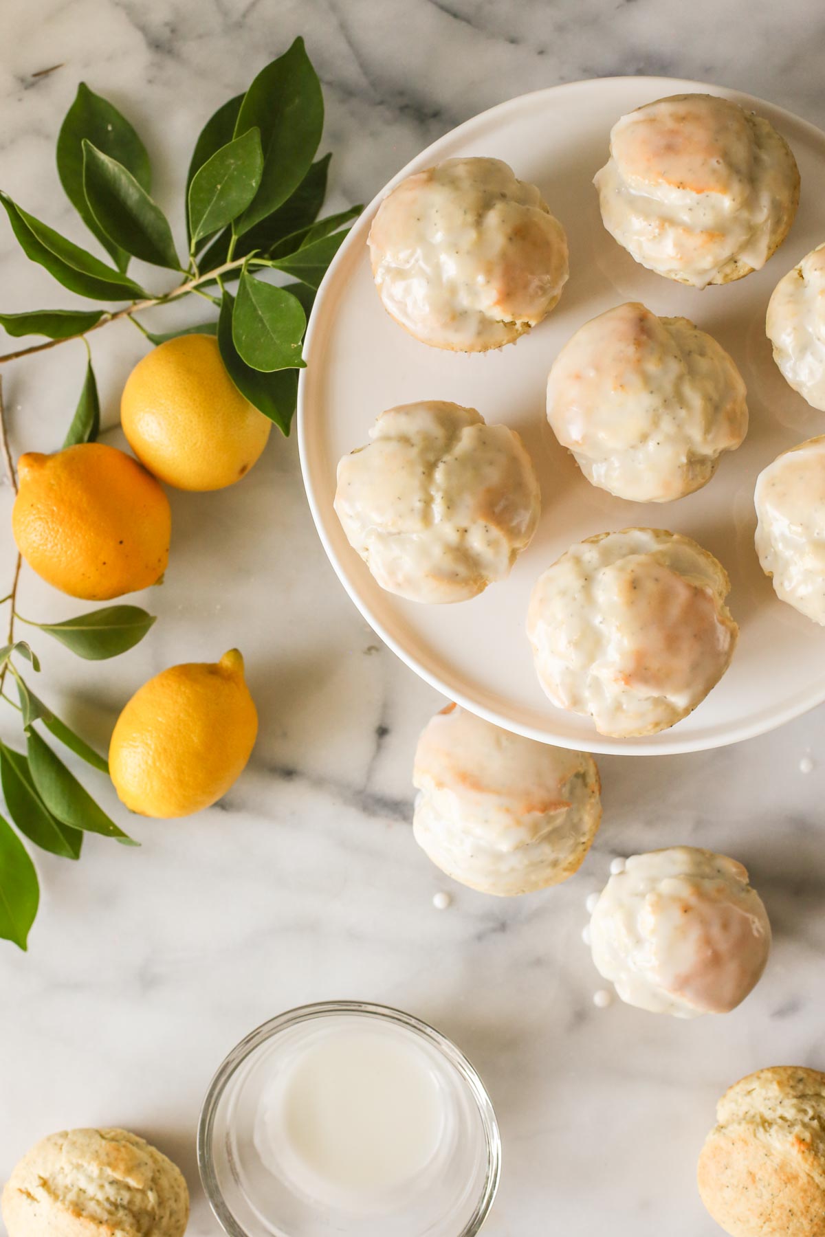 Overhead shot of Glazed Lemon Poppy Seed Muffins on a cake stand with two muffins next to the stand and two unglazed muffins next to a glass bowl of glaze, all on a marble background with a branch from a lemon tree and three whole lemons.  