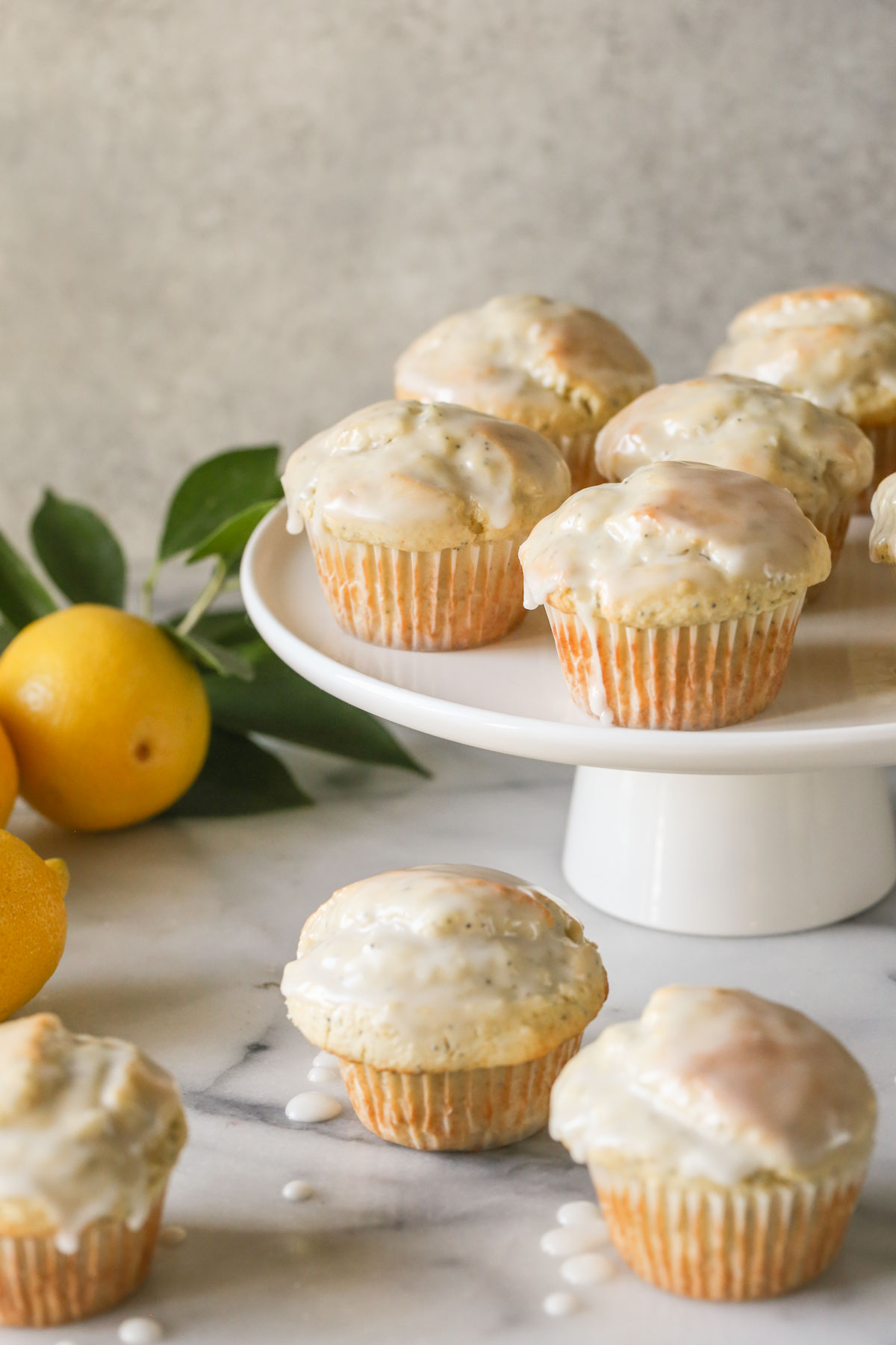 Glazed Lemon Poppy Seed Muffins on a cake stand with three muffins next to the stand, along with a branch from a lemon tree and whole lemons. 