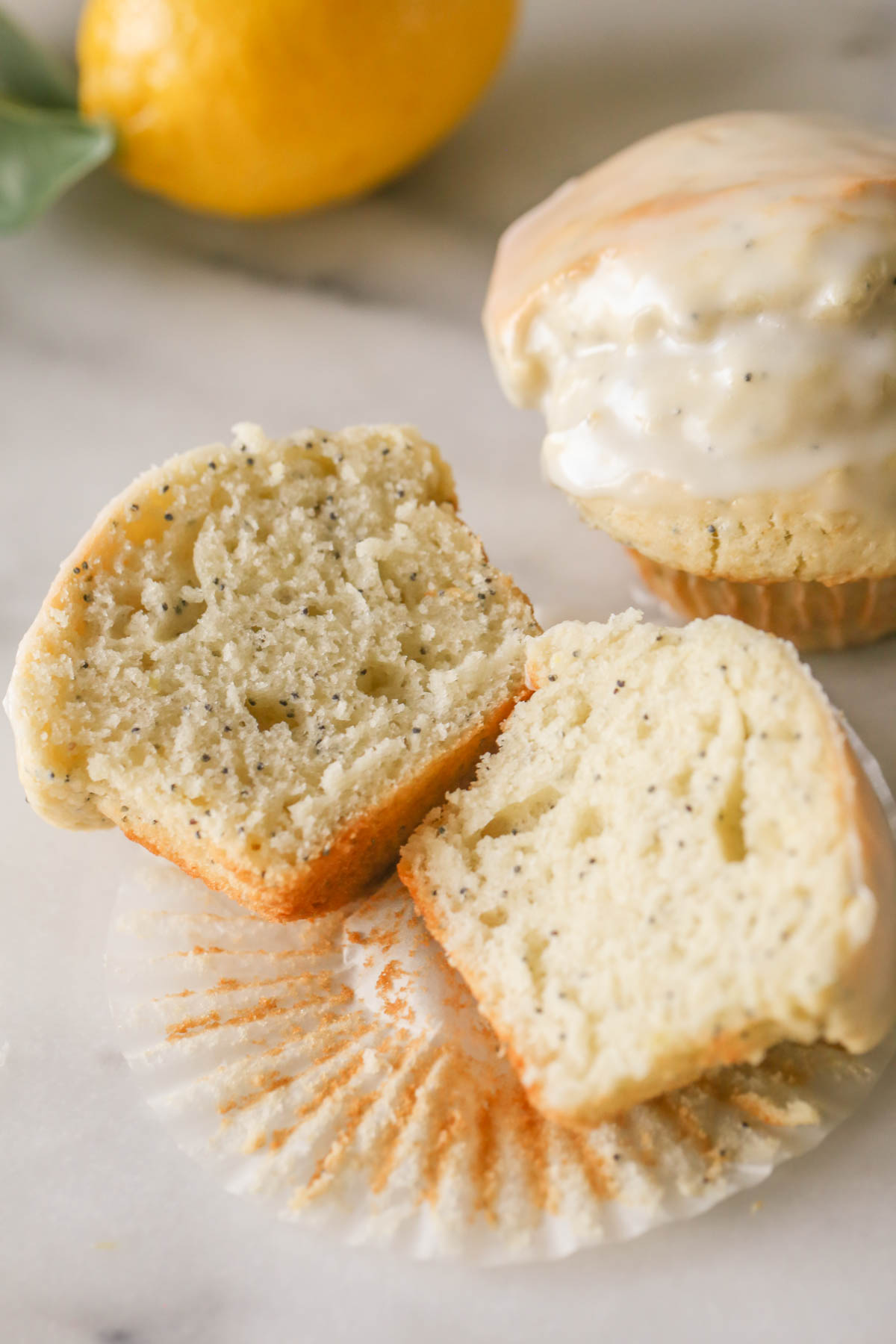 A Glazed Lemon Poppy Seed Muffin sliced in half, placed on the muffin liner, with another whole muffin next to it and a whole lemon in the background. 