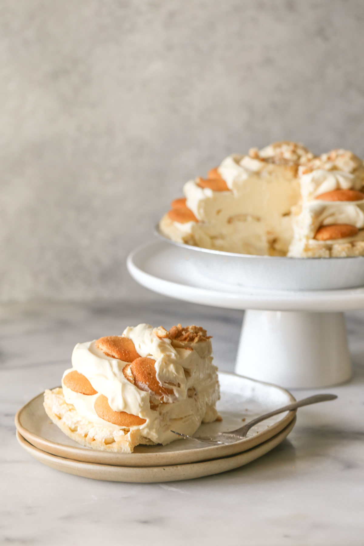 A piece of Mile High Banana Pudding Pie on a plate with a fork, and the rest of the Mile High Banana Pudding Pie on a white cake stand in the background. 