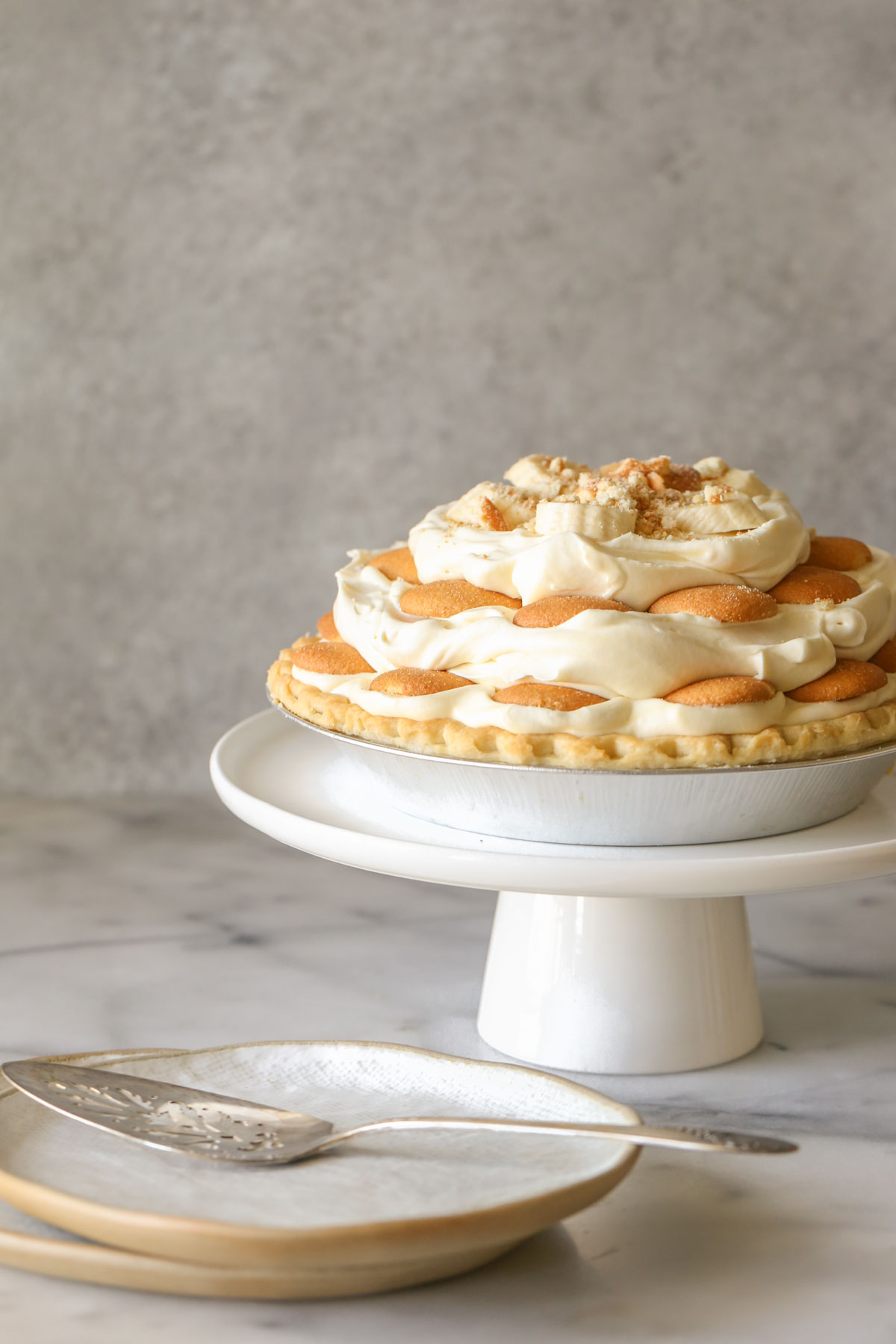 Mile High Banana Pudding Pie on a white cake stand, with two plates and a pie serving utensil next to the cake stand.  