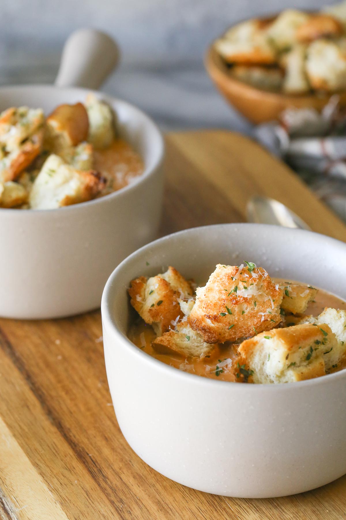 Two bowls of Roasted Vegetable Soup topped with Rustic Buttery Garlic Croutons and fresh Parmesan cheese, sitting on a wood cutting board. 