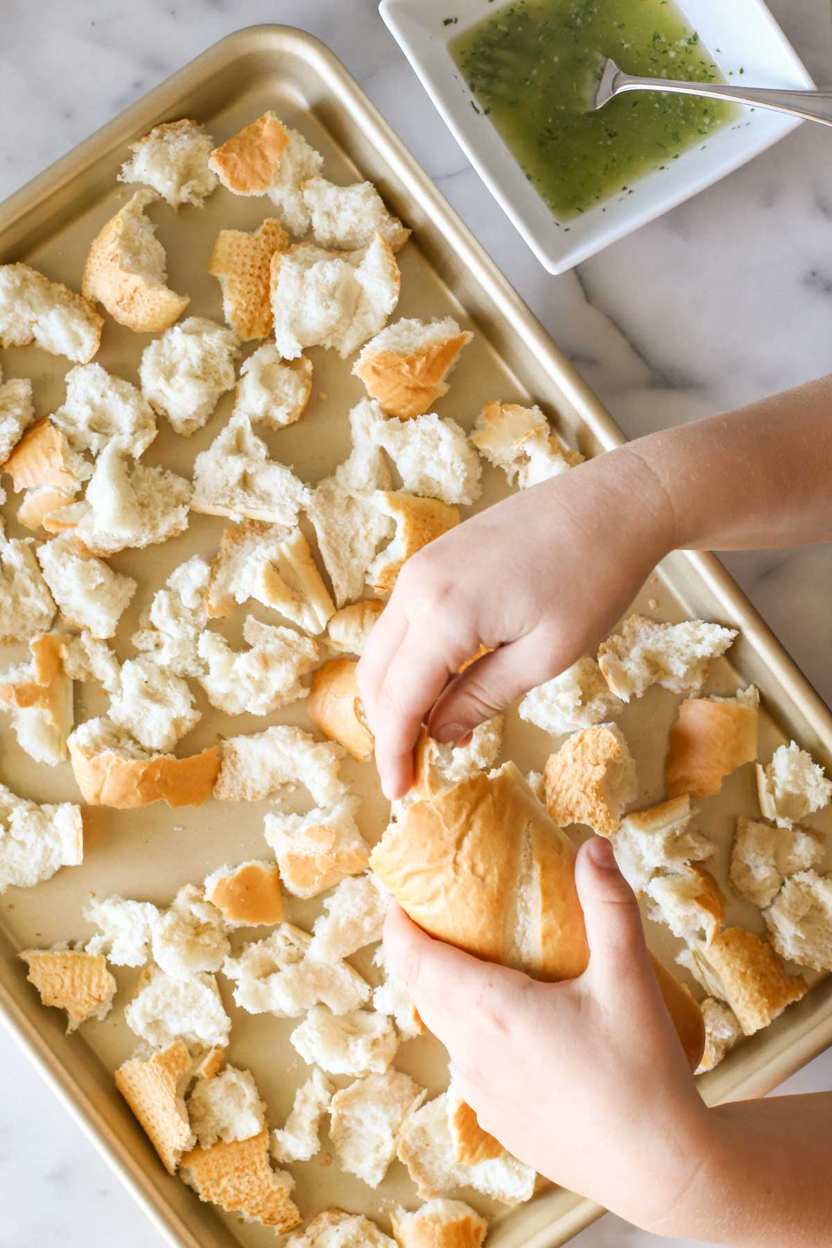 Overhead shot of a hand tearing the French bread into pieces over a baking sheet, with a square bowl of the butter mixture next to the baking sheet. 