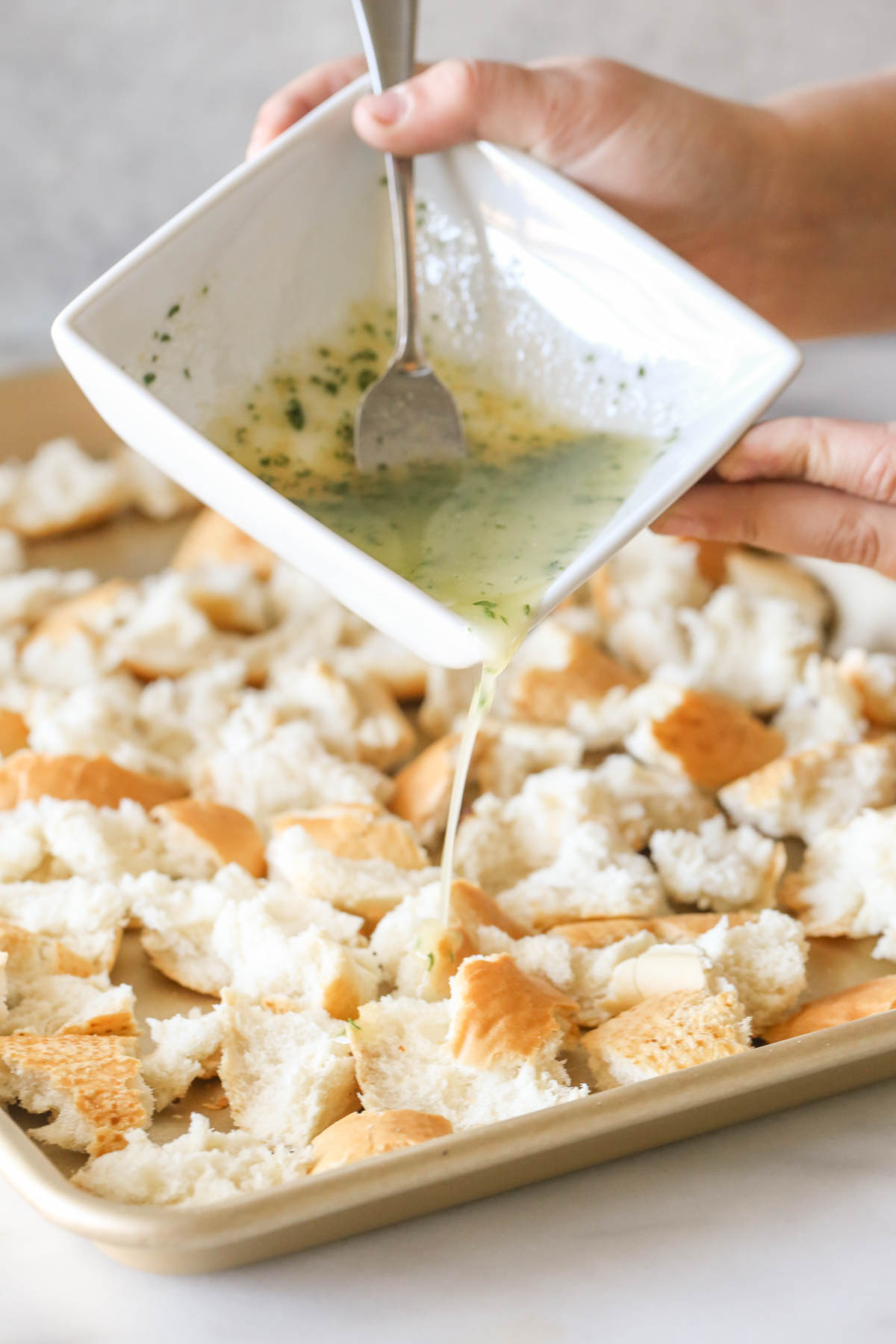 The butter mixture in a square bowl being poured over the French bread pieces on the baking sheet to make Rustic Buttery Garlic Croutons.  