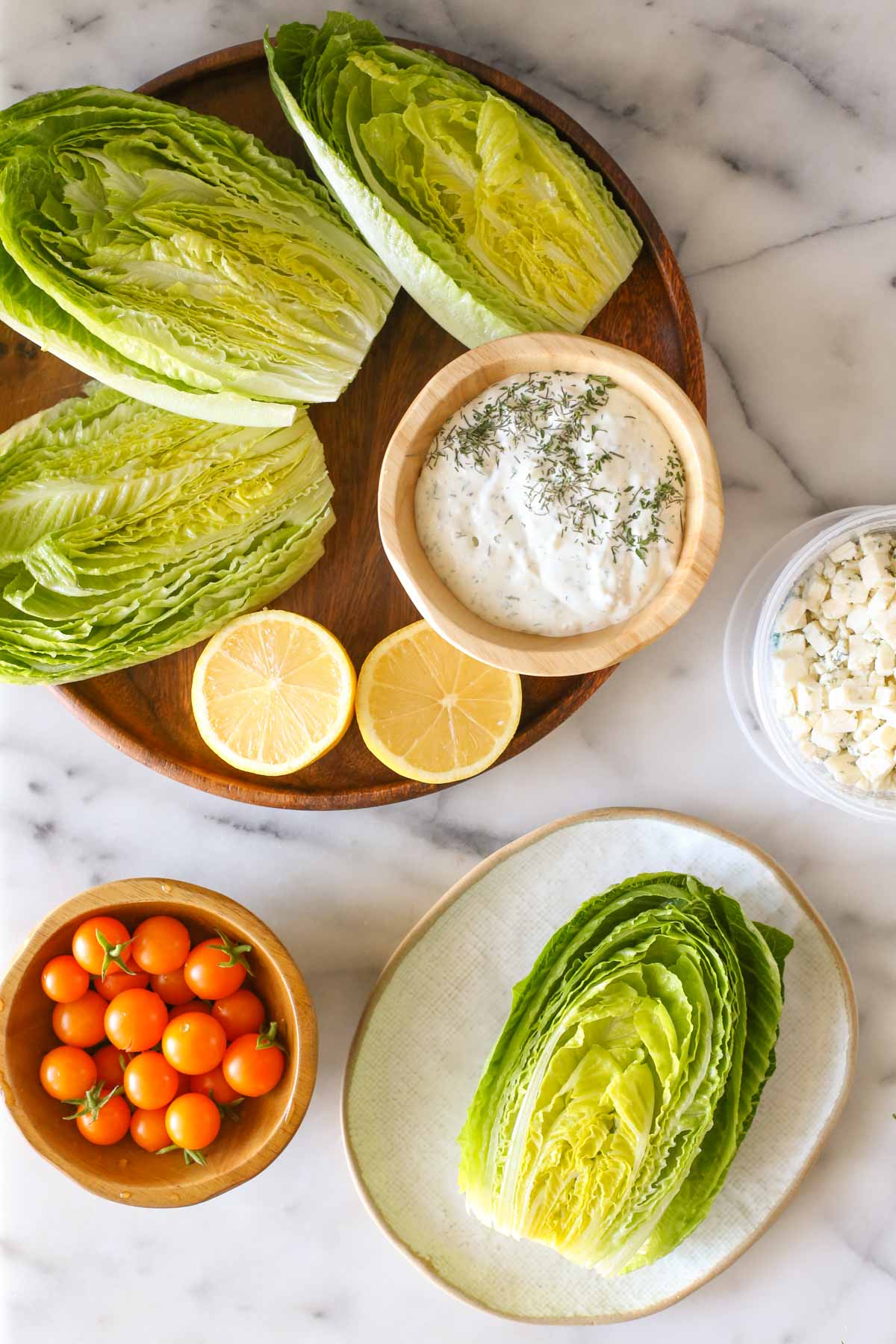 Overhead shot of a wood platter with three romaine lettuce halves, lemon wedges and a wood bowl of the blue cheese dressing on it, sitting next to a wood bowl of Sungold tomatoes, a container of blue cheese and a plate with a romaine lettuce half on it, all on a marble background.  